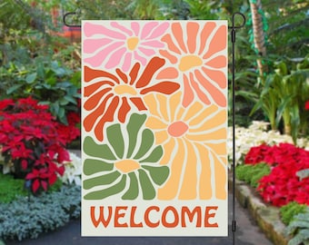 May include: A welcome garden flag with a cream background and a colorful abstract floral design. The flowers are in shades of pink, orange, red, and green. The word "WELCOME" is printed in red at the bottom of the flag.
