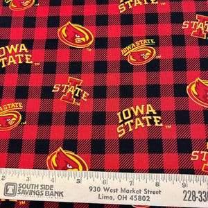 May include: Red and black plaid fabric featuring the Iowa State University logo and the words "IOWA STATE" in gold. The logo is a cardinal bird in a circle. The fabric has a repeating pattern of the logo and text.