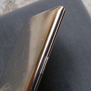 May include: A silver cigarette case with a textured surface. The case is closed and has a slight curve to it.