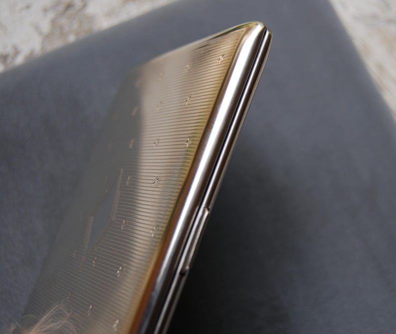 May include: A silver cigarette case with a textured surface. The case is closed and has a slight curve to it.