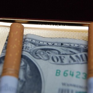 Peut inclure: Deux cigarettes brunes sont plac&eacute;es dans un &eacute;tui &agrave; cigarettes en or avec un billet de dollar en dessous. Le billet de dollar porte les mots "United States of America" et le num&eacute;ro de s&eacute;rie "B 6423".