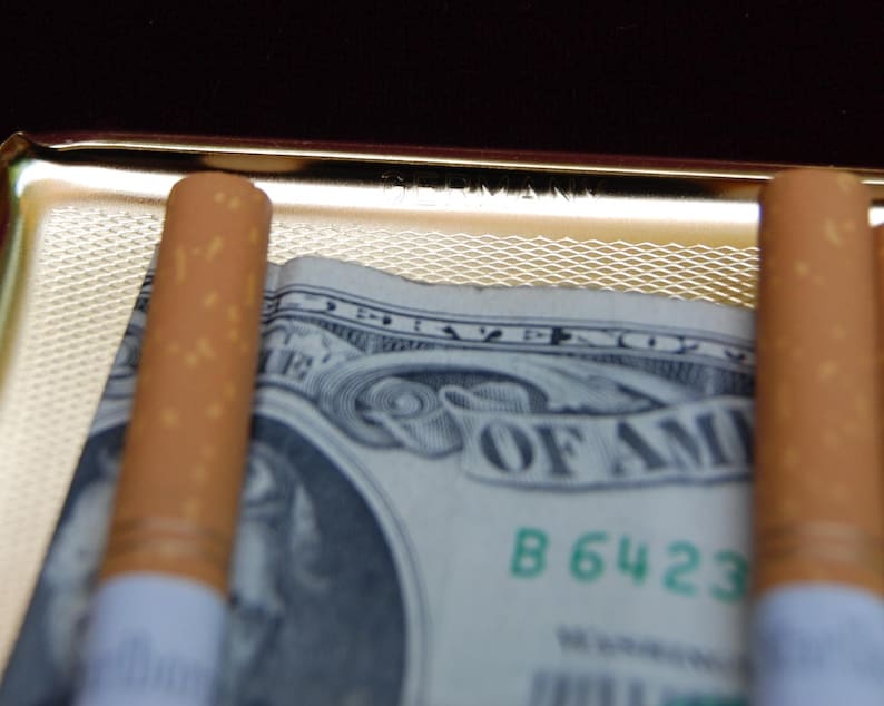 Peut inclure: Deux cigarettes brunes sont plac&eacute;es dans un &eacute;tui &agrave; cigarettes en or avec un billet de dollar en dessous. Le billet de dollar porte les mots "United States of America" et le num&eacute;ro de s&eacute;rie "B 6423".