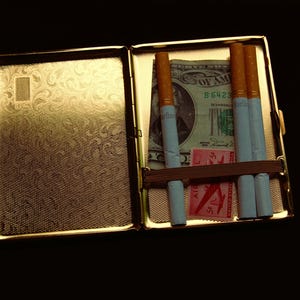 Peut inclure: Un &eacute;tui &agrave; cigarettes en m&eacute;tal dor&eacute; avec un motif. Le bo&icirc;tier est ouvert et contient quatre cigarettes et un billet de dollar. Le billet de dollar porte le texte "United States of America" et "B 6423".