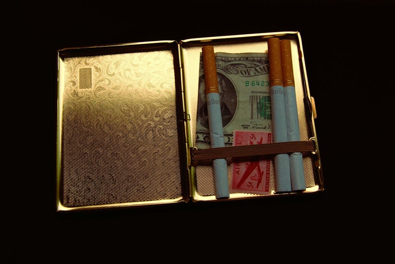 Peut inclure: Un &eacute;tui &agrave; cigarettes en m&eacute;tal dor&eacute; avec un motif. Le bo&icirc;tier est ouvert et contient quatre cigarettes et un billet de dollar. Le billet de dollar porte le texte "United States of America" et "B 6423".
