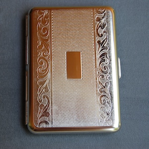 Puede incluir: Una cajetilla de cigarrillos de plata con un diseño ornamentado y un área rectangular en relieve en el centro.