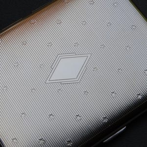May include: A silver cigarette case with a diamond-shaped design and a repeating star pattern.