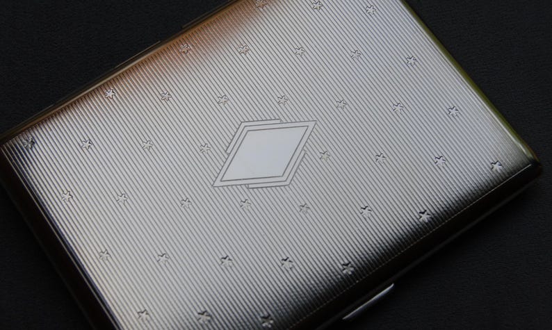 May include: A silver cigarette case with a diamond-shaped design and a repeating star pattern.