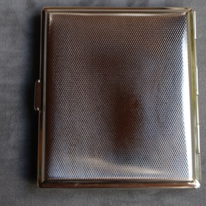 Puede incluir: Un estuche de cigarrillos plateado rectangular con una superficie texturizada con un patr&oacute;n de diamantes. El estuche tiene una tapa con bisagras y un cierre lateral. El estuche est&aacute; cerrado y refleja la luz.