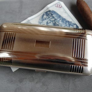 Pode incluir: Um estojo de cigarros de prata com uma superfície texturizada e um design decorativo. O estojo tem uma tampa com dobradiças e um pequeno fecho.