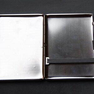 May include: A silver cigarette case with a black elastic band. The case is open and the inside is visible.