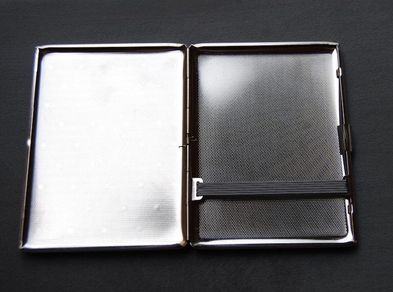 May include: A silver cigarette case with a black elastic band. The case is open and the inside is visible.