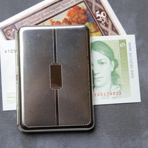 May include: A silver cigarette case with a textured, diagonal pattern and a thin, rectangular, silver band in the center. The case is closed.