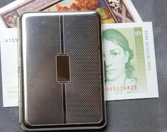 May include: A silver cigarette case with a textured, diagonal pattern and a thin, rectangular, silver band in the center. The case is closed.