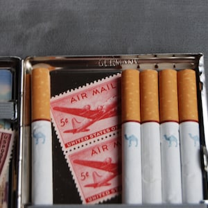 Puede incluir: Cajetilla de cigarrillos abierta, plateada, con cigarrillos y sellos postales Air Mail vintage. Los cigarrillos tienen un logotipo de camello. Los sellos son rojos con una ilustraci&oacute;n de avi&oacute;n. La caja est&aacute; marcada "GERMANY".