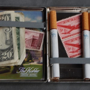 Puede incluir: Un estuche de cigarrillos plateado abierto, que contiene cigarrillos, billetes y sellos. Los cigarrillos tienen tubos blancos y filtros marrones. El estuche tambi&eacute;n contiene un billete de veinte d&oacute;lares, sellos y una postal.