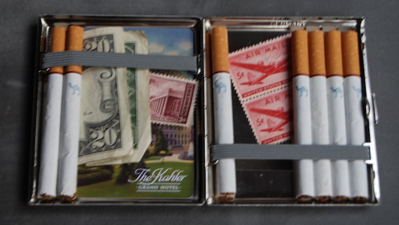 Puede incluir: Un estuche de cigarrillos plateado abierto, que contiene cigarrillos, billetes y sellos. Los cigarrillos tienen tubos blancos y filtros marrones. El estuche tambi&eacute;n contiene un billete de veinte d&oacute;lares, sellos y una postal.