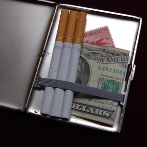 May include: A silver cigarette case containing four cigarettes and a dollar bill. The dollar bill has the text "United States of America" and "B6423084" printed on it. The case also has a pink postage stamp with the text "Air Mail 5&cent;" printed on it.