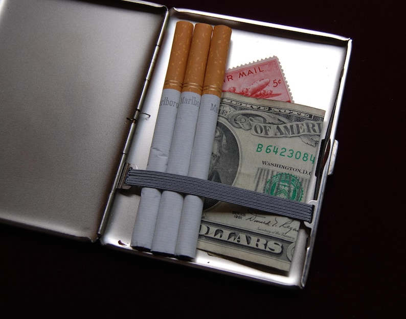 May include: A silver cigarette case containing four cigarettes and a dollar bill. The dollar bill has the text "United States of America" and "B6423084" printed on it. The case also has a pink postage stamp with the text "Air Mail 5&cent;" printed on it.