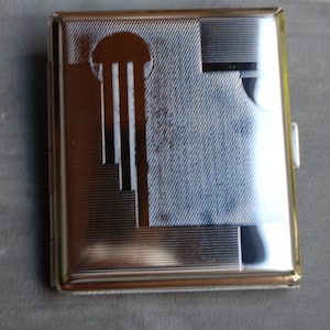 Puede incluir: Un estuche rectangular plateado para cigarrillos con un dise&ntilde;o Art D&eacute;co. El estuche presenta un patr&oacute;n geom&eacute;trico con una serie de l&iacute;neas verticales y una forma semicircular. Tiene una superficie brillante y reflectante, con cierre de broche.