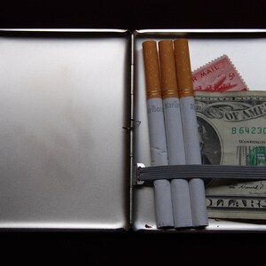 May include: A silver cigarette case with three cigarettes and a dollar bill inside. The dollar bill has the text "United States of America" and "B 6423084" printed on it. The case has a red and white stamp with the text "Air Mail" printed on it.