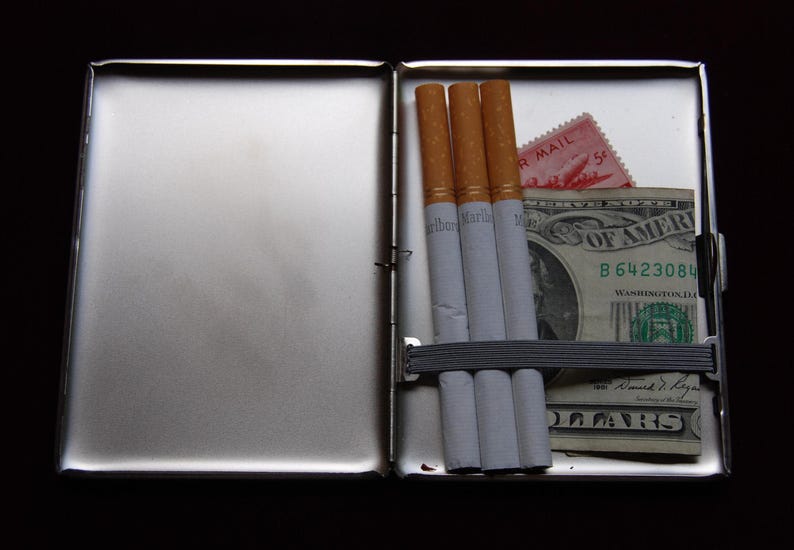 May include: A silver cigarette case with three cigarettes and a dollar bill inside. The dollar bill has the text "United States of America" and "B 6423084" printed on it. The case has a red and white stamp with the text "Air Mail" printed on it.
