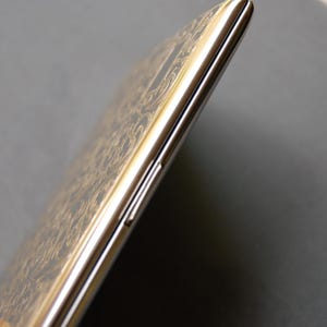 Peut inclure: Un smartphone argent&eacute; et dor&eacute; avec un motif d&eacute;coratif sur le dos. Le t&eacute;l&eacute;phone est pr&eacute;sent&eacute; de c&ocirc;t&eacute;, avec le bouton de volume visible.