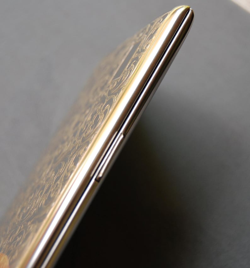 Peut inclure: Un smartphone argent&eacute; et dor&eacute; avec un motif d&eacute;coratif sur le dos. Le t&eacute;l&eacute;phone est pr&eacute;sent&eacute; de c&ocirc;t&eacute;, avec le bouton de volume visible.