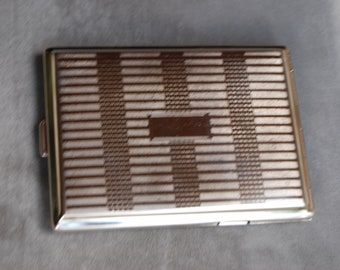 May include: A silver cigarette case with a brown and white geometric pattern. The case has a hinged lid and a small latch.