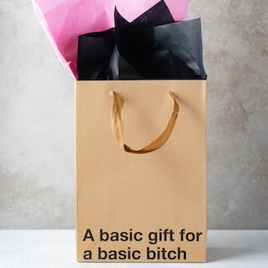 May include: A tan paper gift bag with brown twisted paper handles. The bag features black text printed on the front that reads, A basic gift for a basic bitch. Pink and black tissue paper protrudes from the top opening of the gift bag.