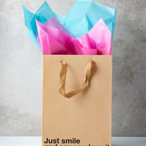 May include: A brown paper gift bag with a gold handle and pink and blue tissue paper inside. The bag has the text "Just smile and say you love it" printed on the bottom.