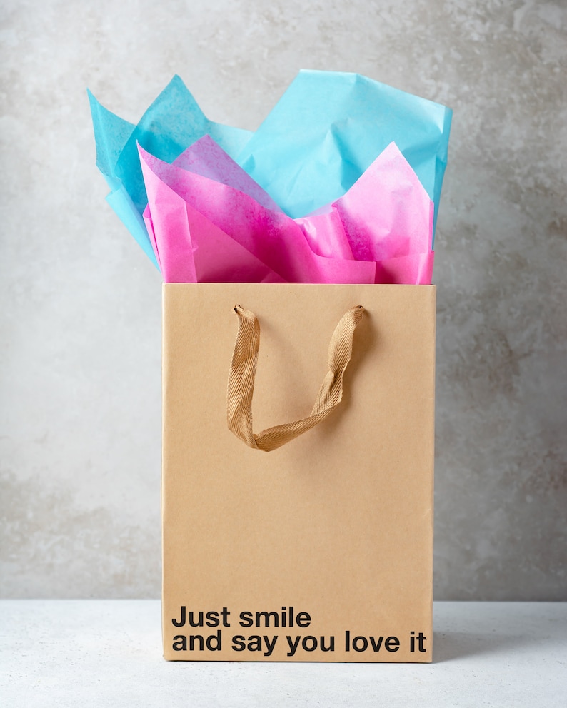 May include: A brown paper gift bag with a gold handle and pink and blue tissue paper inside. The bag has the text "Just smile and say you love it" printed on the bottom.