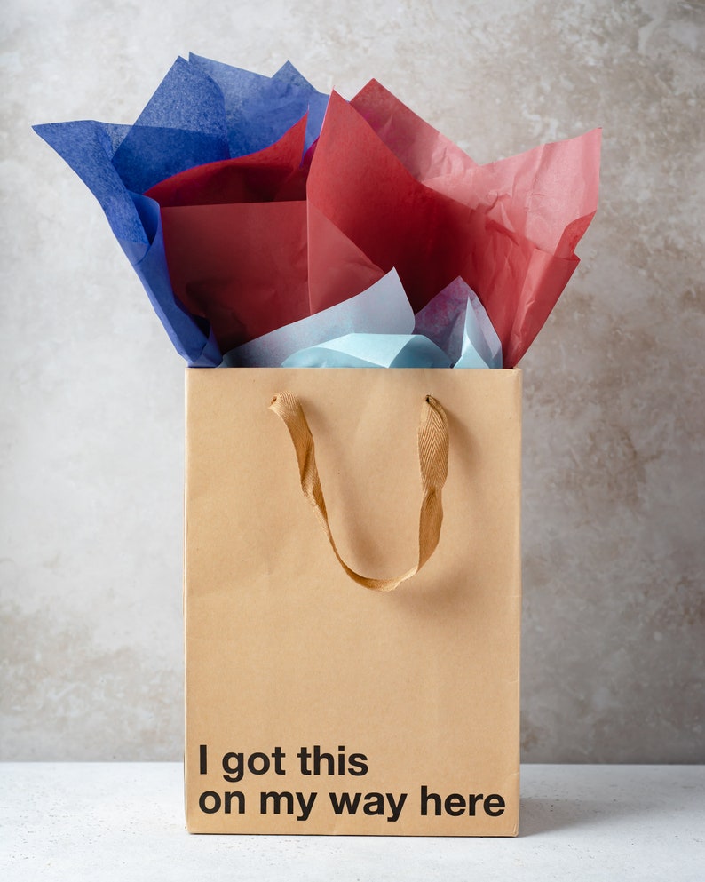 May include: A brown paper gift bag with a brown ribbon handle. The bag is filled with blue, red, and white tissue paper. The bag has the text "I got this on my way here" printed on the front.