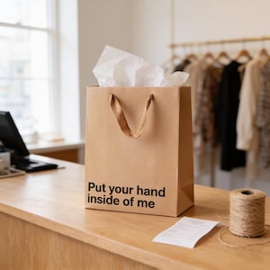 A brown paper shopping bag with the text "Put your hand inside of me" printed on it, placed on a wooden counter in a clothing store.