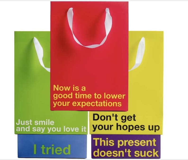 May include: Four colorful gift bags stacked on top of each other. The top bag is red with white handles and the text "Now is a good time to lower your expectations". The second bag is green with white handles and the text "Just smile and say you love it". The third bag is yellow with white handles and the text "Don't get your hopes up". The bottom bag is purple with the text "This present doesn't suck".