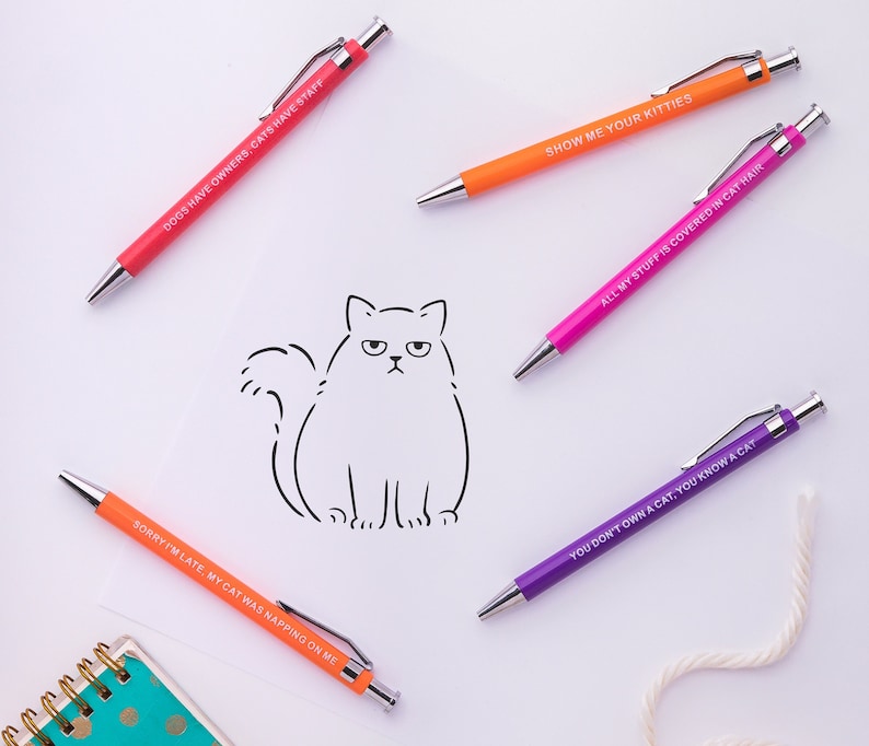 May include: Six colourful pens with humorous cat-themed sayings. The pens are on a white background with a simple line drawing of a cat.
