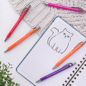 May include: Five colourful pens with humorous cat-themed sayings. The pens are on a white notebook with a line drawing of a cat. The sayings on the pens are: "Dogs have owners, cats have staff", "Show me your kitties", "All my stuff is covered in cat hair", "Sorry I'm late, my cat was napping on me", and "You don't own a cat, you know a cat".