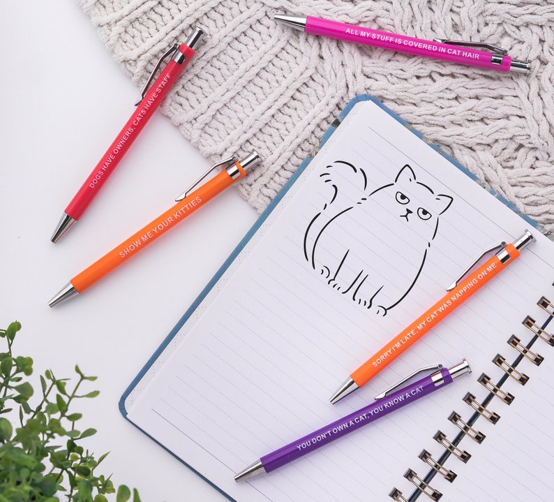 May include: Five colourful pens with humorous cat-themed sayings. The pens are on a white notebook with a line drawing of a cat. The sayings on the pens are: "Dogs have owners, cats have staff", "Show me your kitties", "All my stuff is covered in cat hair", "Sorry I'm late, my cat was napping on me", and "You don't own a cat, you know a cat".