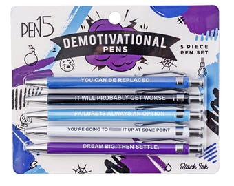 May include: A five-piece set of retractable ballpoint pens with black ink, packaged on a card labeled PEN15 DEMOTIVATIONAL PENS. The pens are blue, black, light blue, white, and purple, each printed with cynical phrases like YOU CAN BE REPLACED and FAILURE IS ALWAYS AN OPTION.