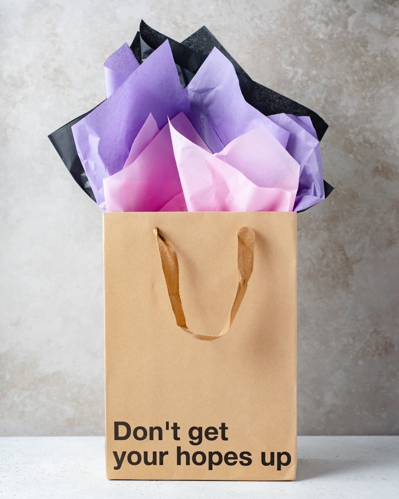 May include: A brown paper gift bag with a black handle and the text "Don't get your hopes up" printed on the front. The bag is filled with pink, purple, and black tissue paper.