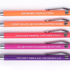 May include: Five colourful pens with humorous cat-themed sayings. The pens are orange, peach, pink, and purple. The sayings are: "Show me your kitties", "Sorry I'm late, my cat was napping on me", "Dogs have owners, cats have staff", "All my stuff is covered in cat hair", and "You don't own a cat, you know a cat".
