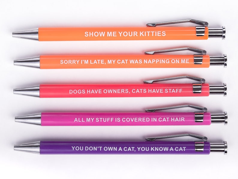 May include: Five colourful pens with humorous cat-themed sayings. The pens are orange, peach, pink, and purple. The sayings are: "Show me your kitties", "Sorry I'm late, my cat was napping on me", "Dogs have owners, cats have staff", "All my stuff is covered in cat hair", and "You don't own a cat, you know a cat".