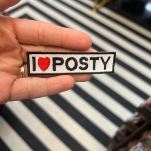 Op de afbeelding: Een rechthoekige patch met de tekst "I ❤️ POSTY" in zwarte letters op een witte achtergrond. Een rood hart vervangt het woord "love". De patch wordt in een hand gehouden.