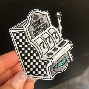 May include: Embroidered patch featuring a slot machine design. The patch is primarily black, white, and gray, with the text "LUCKY COWGIRL" and the number "777" visible. The slot machine has a lever and a checkered pattern.