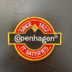 Op de afbeelding: Geborduurde patch met een cirkelvormig ontwerp met de tekst "SINCE 1822" en "IT SATISFIES" in wit op een rode achtergrond. Het woord "Copenhagen" staat in wit op een zwarte rechthoekige banner, met een gele rand.