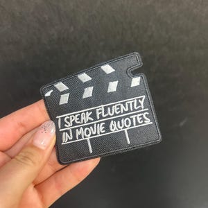 Peut inclure: Un écusson noir en forme de clap de cinéma, avec le texte blanc brodé "I SPEAK FLUENTLY IN MOVIE QUOTES". L'écusson a un aspect texturé et est tenu par une main.
