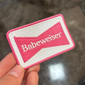 Peut inclure: Écusson brodé avec un motif rose et blanc. L'écusson présente le mot "Babeweiser" en blanc, stylisé en forme de nœud papillon rose, sur un fond blanc et une bordure rose. L'écusson est rectangulaire.