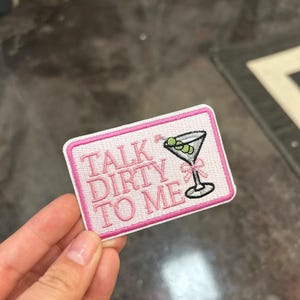 May include: Embroidered patch with the phrase "TALK DIRTY TO ME" in pink lettering. The patch features a martini glass with olives and a pink bow. The patch has a white background with a pink border.