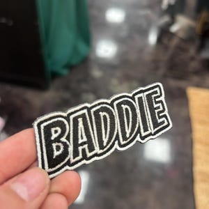 May include: A black and white embroidered patch with the word "BADIE" in bold, capital letters. The patch has a white background with a black border. The patch is held up by a hand.