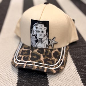 May include: A beige baseball cap featuring a rectangular black and white patch with a portrait of a woman, a silver star-shaped appliqué, and a curved brim with a brown and black leopard print pattern accented by two thin white horizontal stripes.