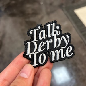 Puede incluir: Un parche bordado en blanco y negro con el texto "Talk Derby to me". El parche tiene un borde negro y letras blancas en una fuente cursiva. El parche se sostiene en una mano.
